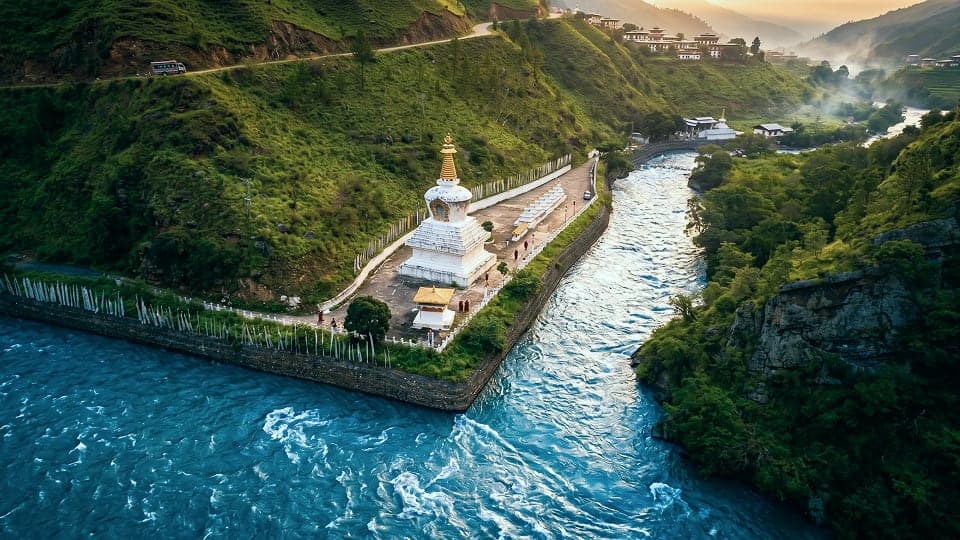 A white chorten with a golden finial standing above a turquoise river bend, prayer flags lining the walkway beside it