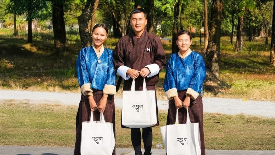 Three young Bhutanese people in traditional dress holding tote bags printed with Dzongkha script, standing outdoors among trees