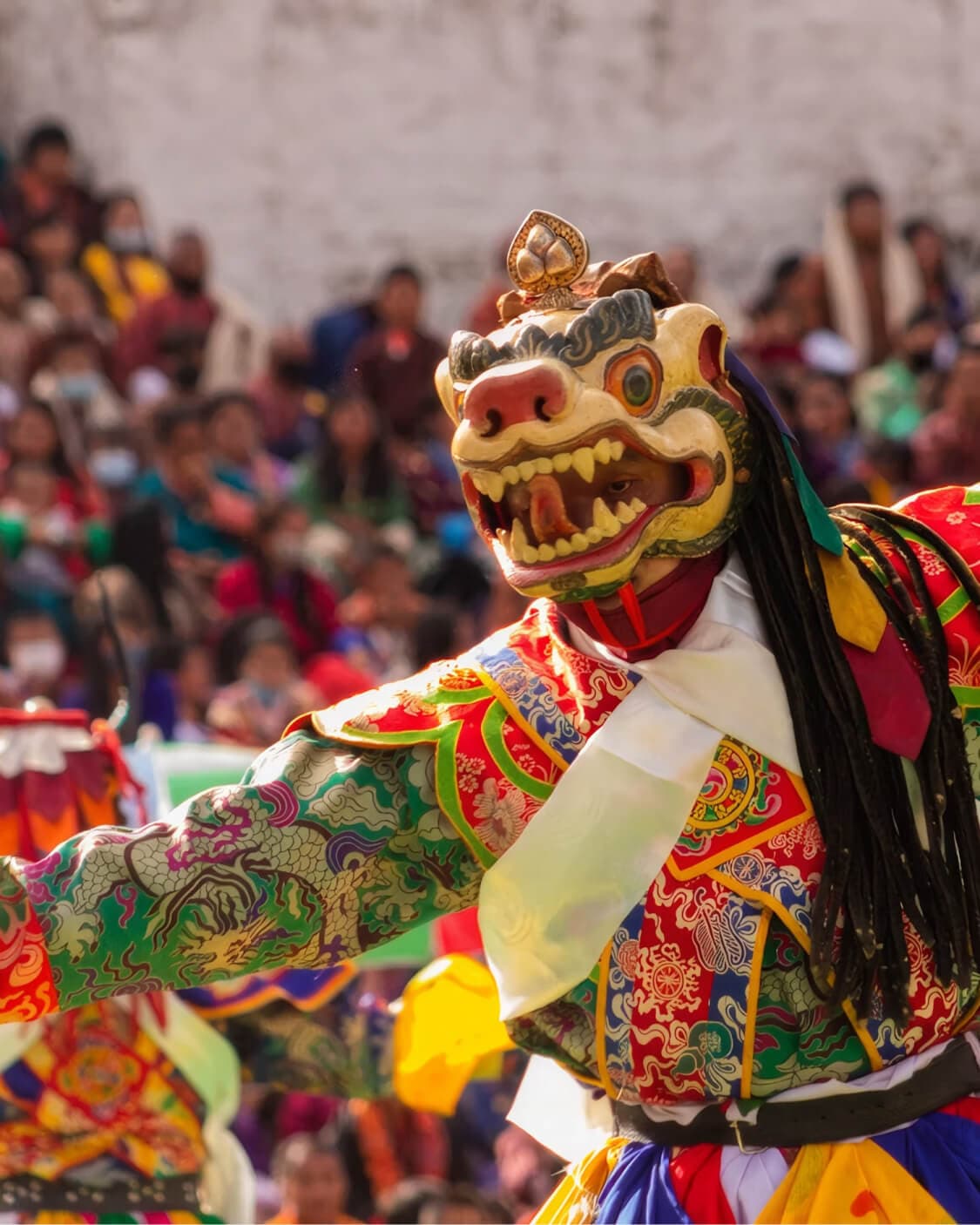 A masked cham dancer in ornate red and gold robes, mid-performance before a crowd.