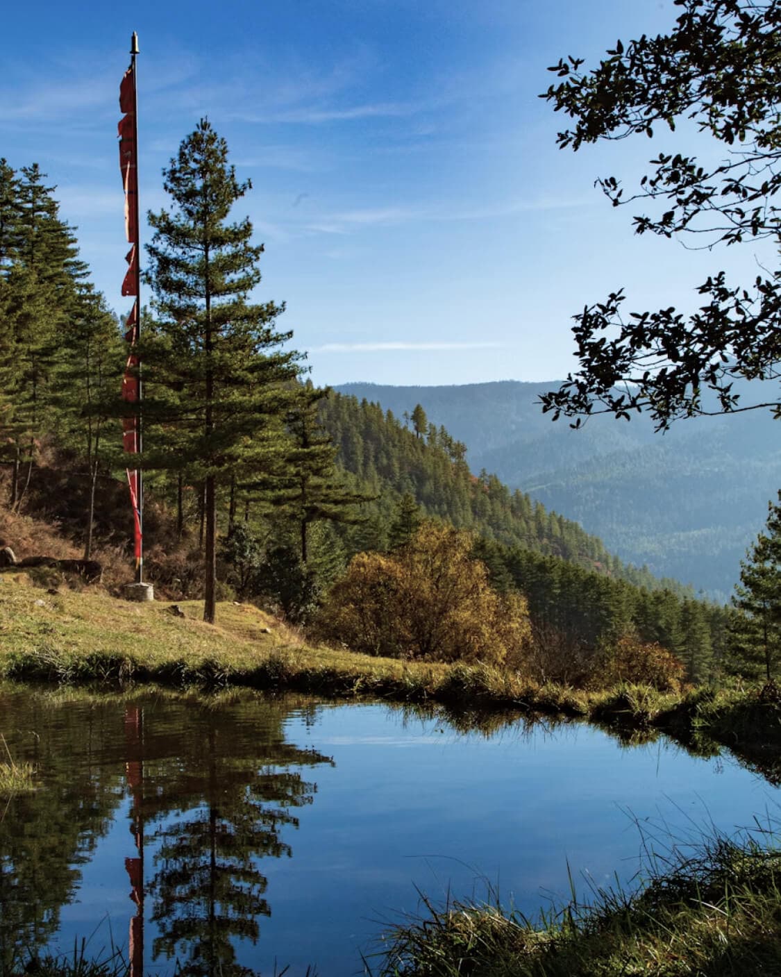 A prayer-flag pole beside a still mountain pond, with pine forest and Himalayan ridgelines reflected in the water.