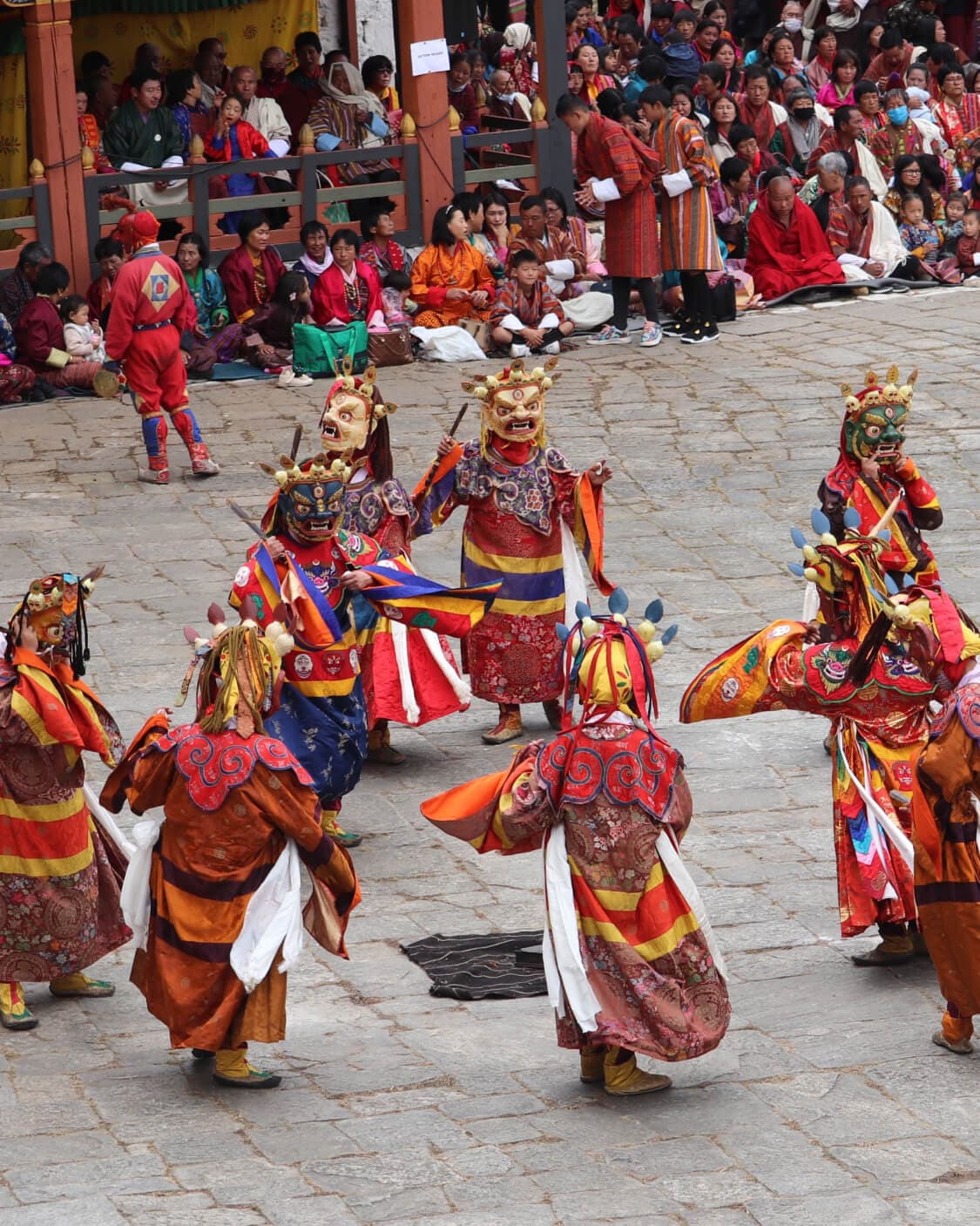 Masked cham dancers performing in a dzong courtyard during a tshechu, watched by a crowd in traditional dress.